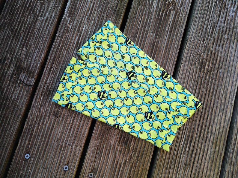 Pu&ograve; includere: Un oggetto di tessuto piegato con un motivo ripetuto di limoni gialli e api nere su sfondo azzurro. Il tessuto &egrave; appoggiato su una superficie di legno invecchiato.