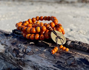 May include: A wooden beaded necklace with a brown cord and a metal charm. The necklace is on a piece of driftwood on a sandy beach.