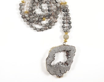 May include: A grey and white stone mala necklace with a large, grey, crystal pendant. The pendant has a hole in the centre and is surrounded by gold accents.
