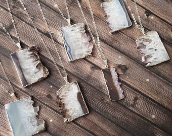 May include: Seven silver necklaces with amethyst crystal pendants. Each pendant is a rectangular slice of amethyst with a natural, rough edge. The amethyst is a light purple colour with white streaks.