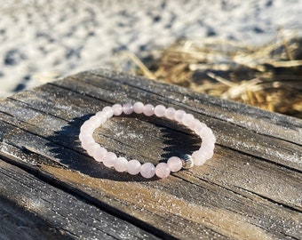 May include: A pink rose quartz beaded bracelet with a silver bead accent. The bracelet is lying on a wooden surface.