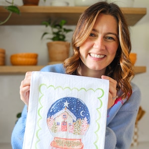 May include: A white kitchen towel with a green scalloped border features a colorful illustration of a snow globe with a small house inside. The snow globe is surrounded by a blue sky with stars and a crescent moon.