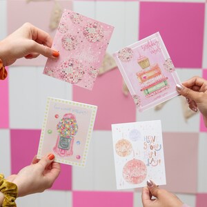 May include: Four pink and white greeting cards with different designs. The cards feature illustrations of disco balls, a gumball machine, a cupcake on a stack of books, and a disco ball with the text "Have a groovy birthday!"