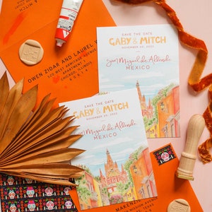 Op de afbeelding: Twee save-the-date-kaarten met een aquarel-illustratie van San Miguel de Allende, Mexico. De kaarten zijn oranje en roze met de tekst "Save the Date" en "Gaby & Mitch" in een vet, oranje lettertype. De tekst "November 20, 2022" en "San Miguel de Allende, Mexico" staat in een kleiner, zwart lettertype. De kaarten worden omringd door andere huwelijkspapieren, waaronder een oranje envelop, een zegellak en een palmblad.