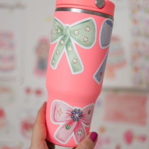 May include: A pink stainless steel tumbler with a lid, decorated with two bow-shaped stickers. One bow is green with white polka dots and the other is pink with rhinestones.