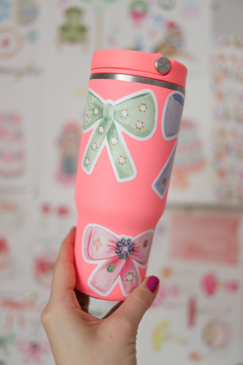 May include: A pink stainless steel tumbler with a lid, decorated with two bow-shaped stickers. One bow is green with white polka dots and the other is pink with rhinestones.