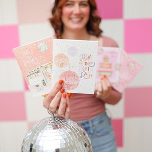 May include: A woman holds up two greeting cards with a disco ball theme. The cards say "Time to Party" and "Have a Groovy Birthday". She is holding a silver disco ball in front of her.