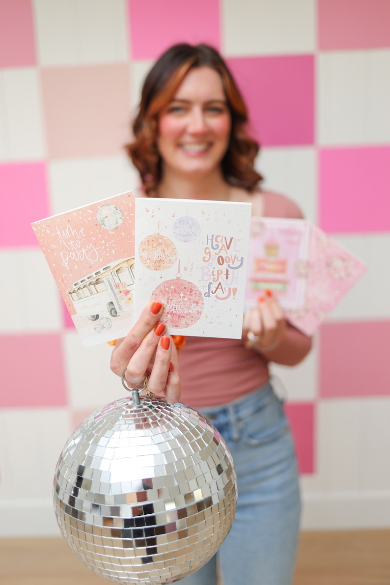 May include: A woman holds up two greeting cards with a disco ball theme. The cards say "Time to Party" and "Have a Groovy Birthday". She is holding a silver disco ball in front of her.