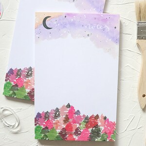 May include: White notepad with a watercolor design featuring a forest at the bottom and a night sky with a crescent moon and stars at the top. The word "notes" is written in cursive. Gold scissors and paint brushes are also in the image.
