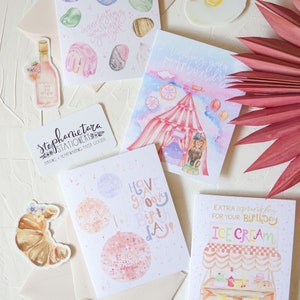 May include: A set of five watercolor greeting cards with various designs. The cards feature a crystal design, a birthday circus tent, a disco ball, an ice cream stand, and a croissant. The cards are all white with pink, blue, and yellow accents. The cards are perfect for sending birthday wishes or other special occasions.
