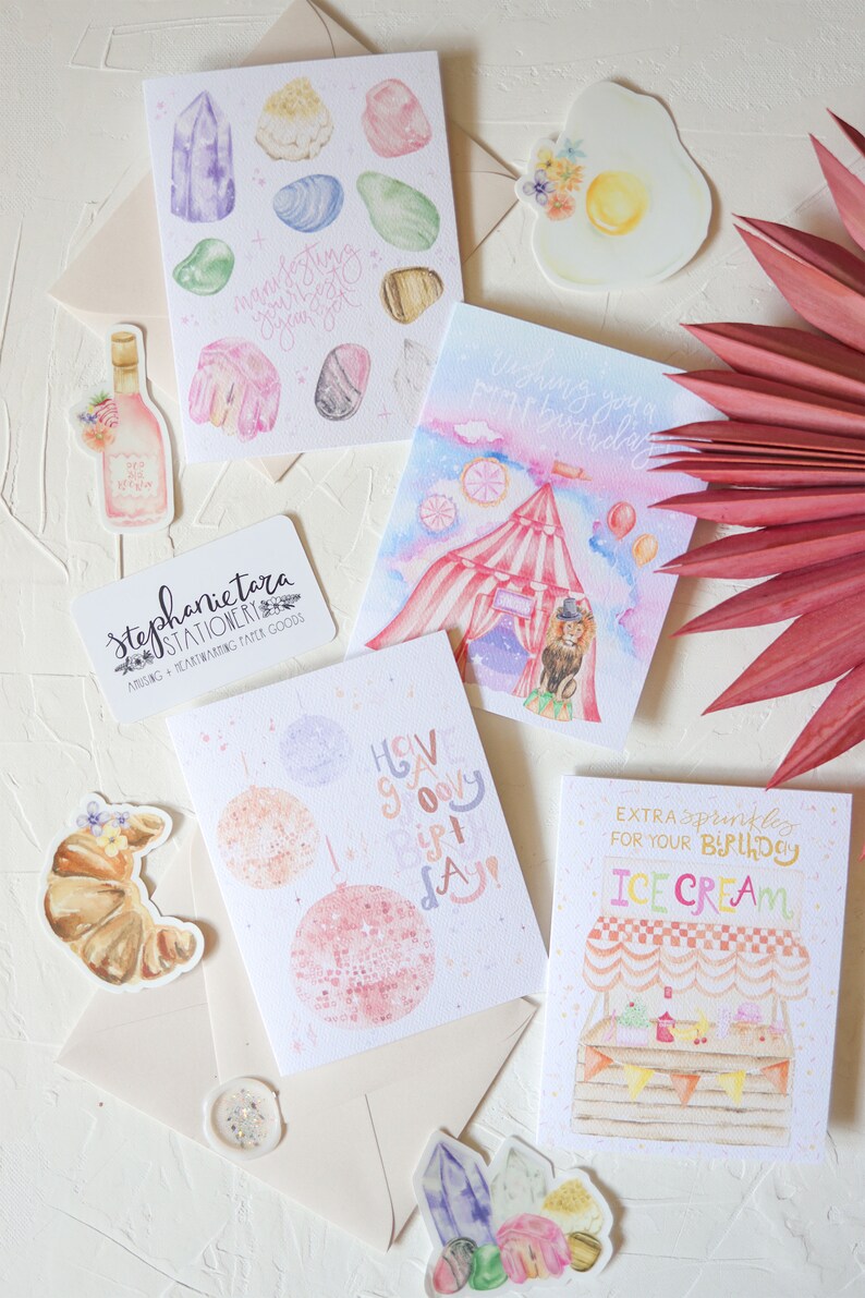 May include: A set of five watercolor greeting cards with various designs. The cards feature a crystal design, a birthday circus tent, a disco ball, an ice cream stand, and a croissant. The cards are all white with pink, blue, and yellow accents. The cards are perfect for sending birthday wishes or other special occasions.