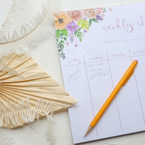 May include: A floral weekly planner with a yellow pencil on top. The planner features a floral design with pink, purple, and orange flowers. The planner has a section for each day of the week, starting with Sunday and ending with Friday. The planner has the words "weekly things" written at the top.