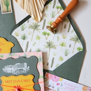 May include: A green envelope with a white and green patterned liner. The envelope is sealed with a wax seal and a wooden stamp. The envelope is addressed to Anakaren and Joseph. The envelope is decorated with a pink and green floral pattern. The envelope is tied with a yellow ribbon and a pink tassel. The envelope is decorated with a yellow tag that says "Meet us in Cabo!" and a website address. The envelope is decorated with a cactus stamp.