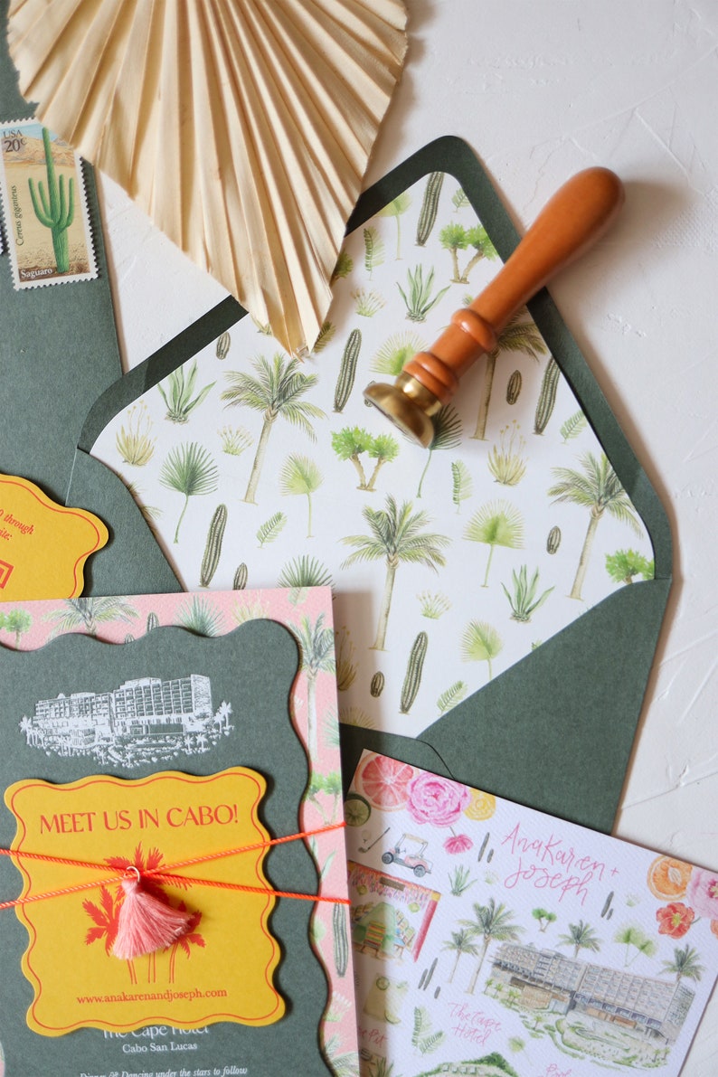 May include: A green envelope with a white and green patterned liner. The envelope is sealed with a wax seal and a wooden stamp. The envelope is addressed to Anakaren and Joseph. The envelope is decorated with a pink and green floral pattern. The envelope is tied with a yellow ribbon and a pink tassel. The envelope is decorated with a yellow tag that says "Meet us in Cabo!" and a website address. The envelope is decorated with a cactus stamp.