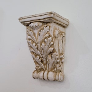 May include: A white plaster wall shelf with an ornate leaf design. The shelf is in the shape of a bracket and has a slightly distressed finish.