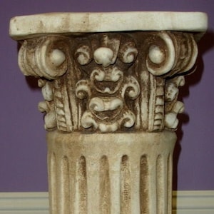 May include: A white, decorative, fluted column pedestal with a detailed, ornate top. The pedestal is made of plaster or ceramic and has a distressed, aged look.