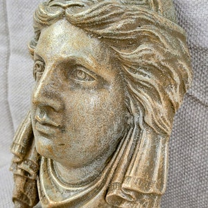 May include: A gold-toned plaster bust of a woman's face with a detailed hairstyle and a draped fabric around the neck and shoulders.