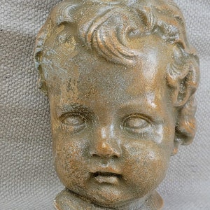 Puede incluir: Una escultura de cerámica de la cabeza de un niño con un acabado desgastado y envejecido. La escultura está pintada en tonos marrones y dorados, y la cara del niño tiene una expresión dulce e inocente.
