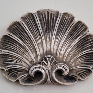 May include: A decorative wall sculpture in the shape of a seashell with a white and gray painted finish. The shell has a detailed, textured design and a scalloped edge.