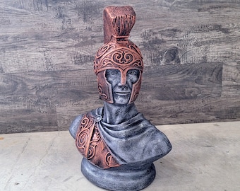 May include: A gray and copper colored bust sculpture of a warrior wearing a helmet with intricate designs. The sculpture is on a gray background.