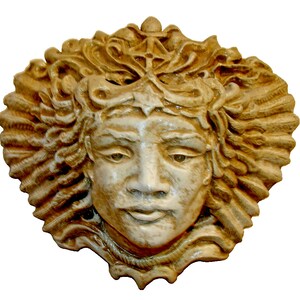 May include: A ceramic wall sculpture of a face with a crown of stylised hair. The face is painted in shades of beige and brown, with a subtle sheen. The sculpture is a decorative piece for a home or garden.