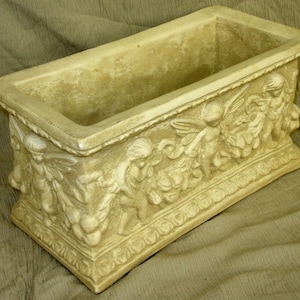 Könnte beinhalten: Ein rechteckiger Pflanzkasten aus hellbeigefarbenem Beton mit einem dekorativen Design aus Cherubim und floralen Motiven am oberen Rand.