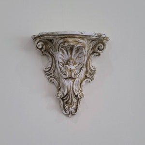 May include: White and gold ornate wall shelf with a decorative design. The shelf is made of plaster or resin and has a distressed finish.