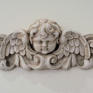 May include: A white plaster cherub with wings, the cherub has a sweet expression and is looking down. The cherub is attached to a decorative scrollwork design.