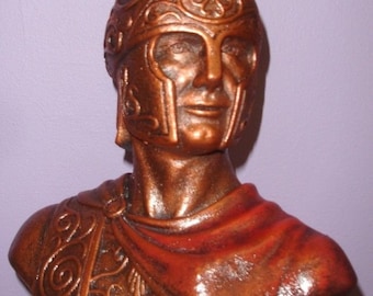 May include: A bronze bust of a Roman soldier wearing a helmet with a plume. The bust is detailed with intricate carvings and a red cloak.