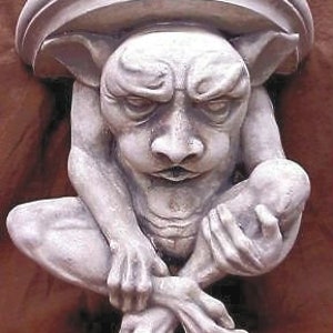 Könnte beinhalten: Eine weiße Steingargouille-Skulptur mit einem detaillierten Gesicht und einem finsteren Ausdruck. Die Gargouille sitzt mit gekreuzten Beinen und ineinander verschränkten Händen. Die Skulptur hat eine flache Oberseite, die als Regal verwendet werden könnte.
