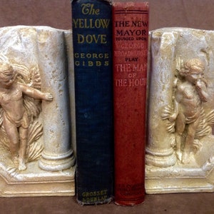 Könnte beinhalten: Ein Paar Buchstützen aus weißem Gips mit goldenen Akzenten. Jede Buchstütze zeigt eine Cherubinskulptur, die eine Säule hält. Die Buchstützen halten zwei Bücher, eines mit einem blauen Einband und dem Titel "The Yellow Dove" von George Gibbs, und das andere mit einem roten Einband und dem Titel "The New Mayor Founded Upon George Broadhurst Play The Man Of The Hou".