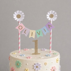 May include: A decorated cake with a banner that reads "OLIVIA" and a gold glitter "1" cake topper. The cake is adorned with pastel-colored flowers and daisy toppers on pink and white striped straws. A festive dessert for a first birthday.