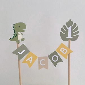 May include: A cake topper featuring a green dinosaur and a monstera leaf with the number 1, connected by a string of pennant flags spelling the name JACOB in white letters. The flags alternate between olive green, mustard yellow, and beige colors.
