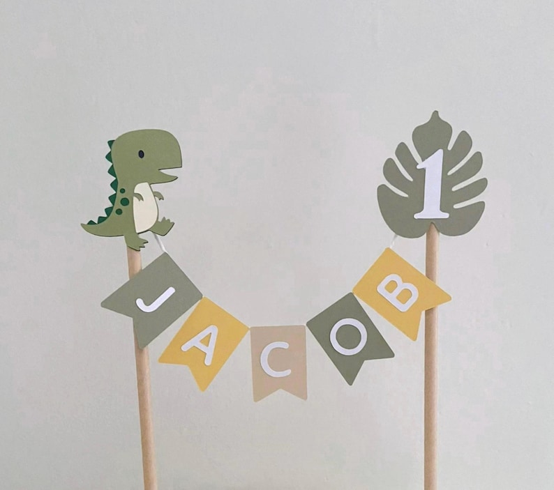 May include: A cake topper featuring a green dinosaur and a monstera leaf with the number 1, connected by a string of pennant flags spelling the name JACOB in white letters. The flags alternate between olive green, mustard yellow, and beige colors.