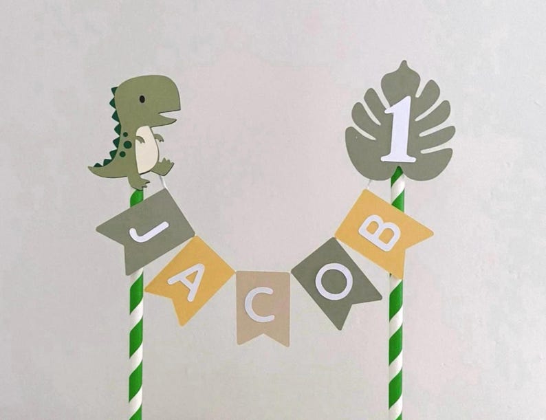 May include: A cake topper featuring two green and white striped paper straws connected by a bunting banner that spells the name JACOB. The banner consists of alternating olive green, yellow, and beige flags. A green dinosaur and a monstera leaf with the number 1 are attached to the top of the straws.