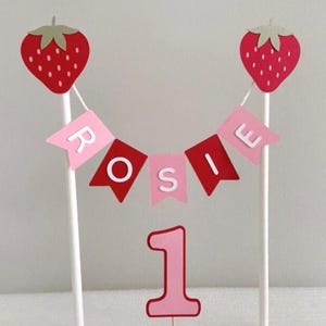 Puede incluir: Un conjunto de decoraci&oacute;n para tartas con una fresa roja a cada lado, una pancarta con la palabra "ROSIE" y el n&uacute;mero "1". Las fresas tienen copas verdes y la pancarta tiene banderas rosas y rojas. El n&uacute;mero "1" es rosa con un contorno rojo.