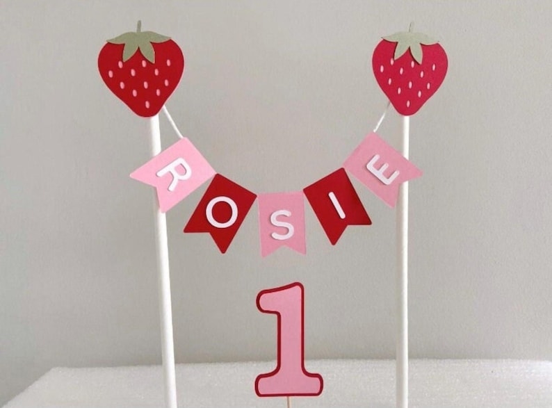 Puede incluir: Un conjunto de decoraci&oacute;n para tartas con una fresa roja a cada lado, una pancarta con la palabra "ROSIE" y el n&uacute;mero "1". Las fresas tienen copas verdes y la pancarta tiene banderas rosas y rojas. El n&uacute;mero "1" es rosa con un contorno rojo.