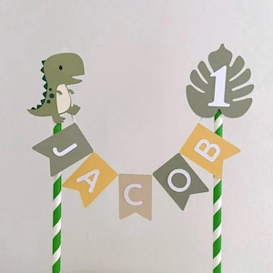 May include: A cake topper featuring two green and white striped paper straws connected by a bunting banner that spells the name JACOB in white letters on alternating olive green, yellow, and tan flags. A green dinosaur and a monstera leaf with the number 1.