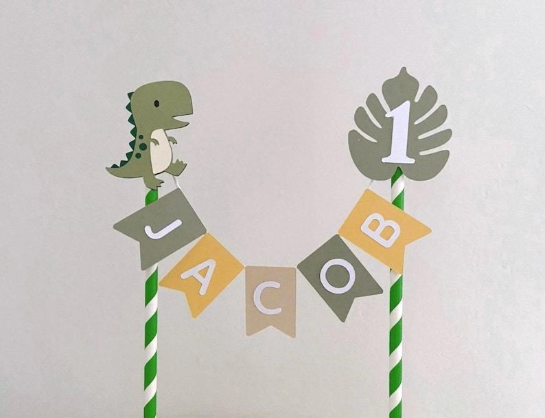 May include: A cake topper featuring two green and white striped paper straws connected by a bunting banner that spells the name JACOB in white letters on alternating olive green, yellow, and tan flags. A green dinosaur and a monstera leaf with the number 1.