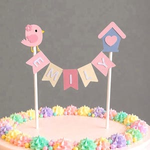 May include: A pink frosted cake decorated with pastel-coloured frosting and a banner that reads "EMILY". The cake is topped with a pink bird and a blue birdhouse with a heart.