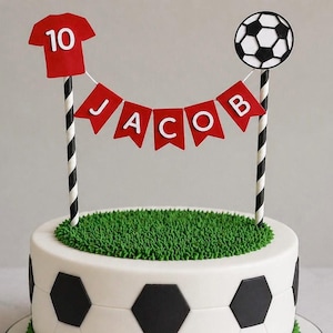 May include: A soccer-themed cake with a white base decorated with black soccer ball shapes. The top features green frosting resembling grass, a red jersey with the number 10, a soccer ball, and a banner that spells "JACOB".