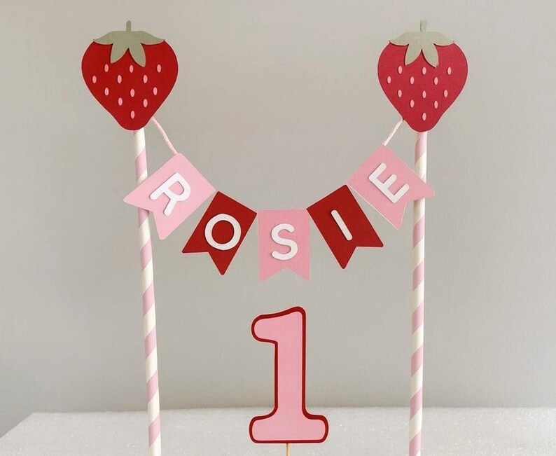 Puede incluir: Un adorno para tarta con tem&aacute;tica de fresas con el nombre "ROSIE" en una pancarta. El adorno incluye dos recortes de fresas rojas, banderines rosas y rojos, el n&uacute;mero "1" rosa y pajitas a rayas rosas y blancas.