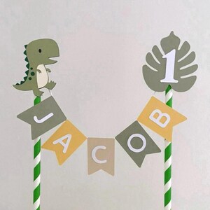 May include: A cake topper featuring two green and white striped paper straws connected by a bunting banner that spells the name JACOB in white letters on alternating olive green, yellow, and tan flags, topped with a dinosaur and a leaf with a number 1.