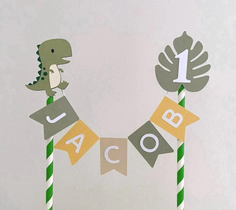 May include: A cake topper featuring two green and white striped paper straws connected by a bunting banner that spells the name JACOB in white letters on alternating olive green, yellow, and tan flags, topped with a dinosaur and a leaf with a number 1.