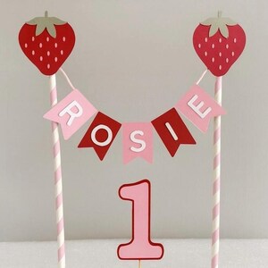 Puede incluir: Adorno para tarta con dos fresas rojas y una pancarta con la palabra "ROSIE". El n&uacute;mero "1" est&aacute; debajo. Las fresas est&aacute;n sobre pajitas a rayas rosas y blancas, ideal para un primer cumplea&ntilde;os.