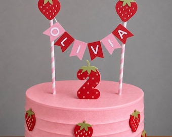 May include: A pink cake topper set featuring a banner with the name OLIVIA in white letters on alternating pink and red pennants, two red strawberry toppers on striped pink and white sticks, and a red strawberry-shaped number two topper.