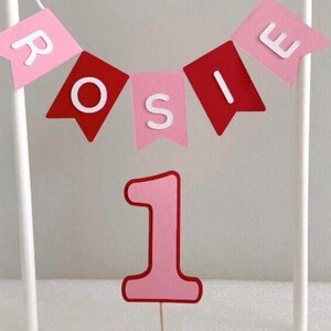 Puede incluir: Adorno para tarta rosa y rojo con el nombre "Rosie" en banderines. Un gran n&uacute;mero "1" debajo, para un primer cumplea&ntilde;os. El adorno est&aacute; sostenido por palitos blancos.