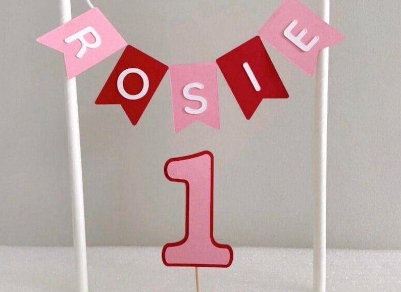 Puede incluir: Adorno para tarta rosa y rojo con el nombre "Rosie" en banderines. Un gran n&uacute;mero "1" debajo, para un primer cumplea&ntilde;os. El adorno est&aacute; sostenido por palitos blancos.