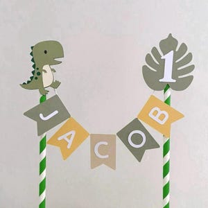 May include: A cake topper featuring two green and white striped paper straws connected by a bunting banner that spells the name JACOB in white letters on alternating olive green and mustard yellow flags, with a green dinosaur and a number 1.