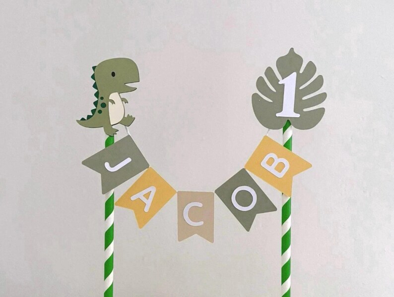 May include: A cake topper featuring two green and white striped paper straws connected by a bunting banner that spells the name JACOB in white letters on alternating olive green and mustard yellow flags, with a green dinosaur and a number 1.